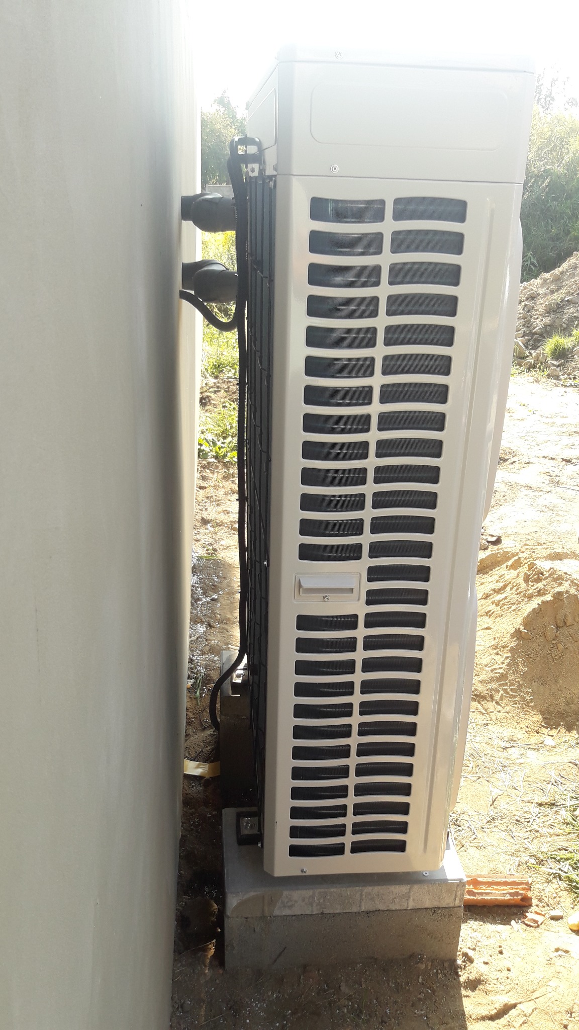 Boczny widok jednostki zewnętrznej klimatyzacji, zamontowanej na betonowym fundamencie przy ścianie budynku, z widocznymi przewodami i otoczeniem placu budowy.