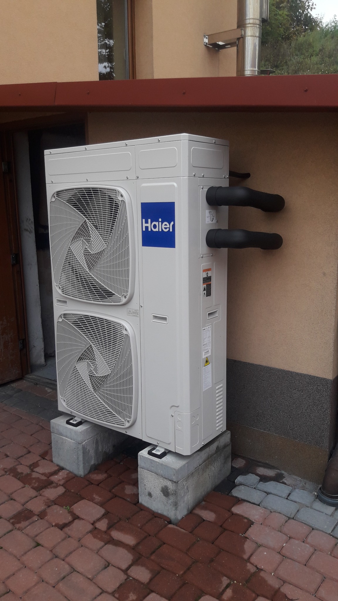 Zewnętrzna jednostka dwuwentylatorowa klimatyzatora Haier, umieszczona na betonowych blokach na ceglanym chodniku, z widocznymi rurami i ścianą budynku w tle.