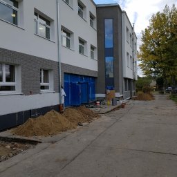 Etna Plus Sp. z o.o. - Budynek w trakcie remontu elewacji i wymiany stolarki okiennej, widoczne okna z białymi ramami, świeżo otynkowana ściana, zabezpieczone wejście i hałda piachu.