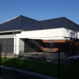 Nowoczesny dom jednorodzinny z szarą elewacją, ciemnym dachem, garażem z roletami zewnętrznymi i drewnopodobną okładziną, widziany zza ogrodzenia z siatki.