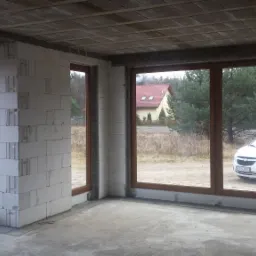 Surowy stan budynku w trakcie budowy z zamontowanymi oknami z brązowymi ramami, widok na zewnątrz na samochód firmy i okoliczny krajobraz.