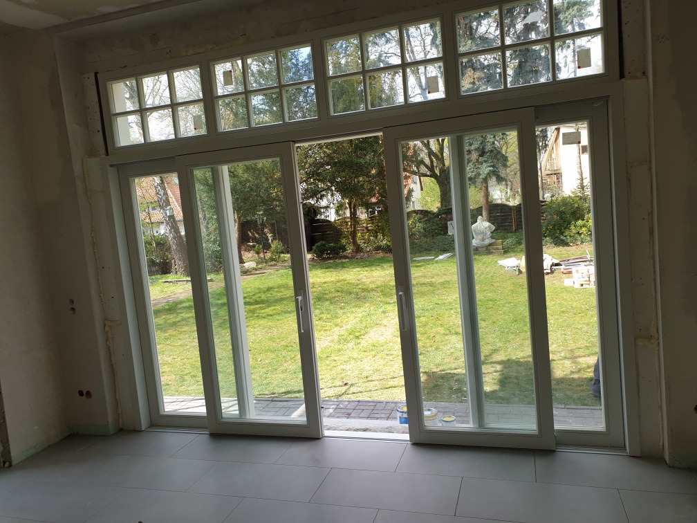 Wnętrze z dużym, przesuwnym oknem z widokiem na zielony ogród, w trakcie montażu, widoczne niedokończone ściany i przygotowane materiały.