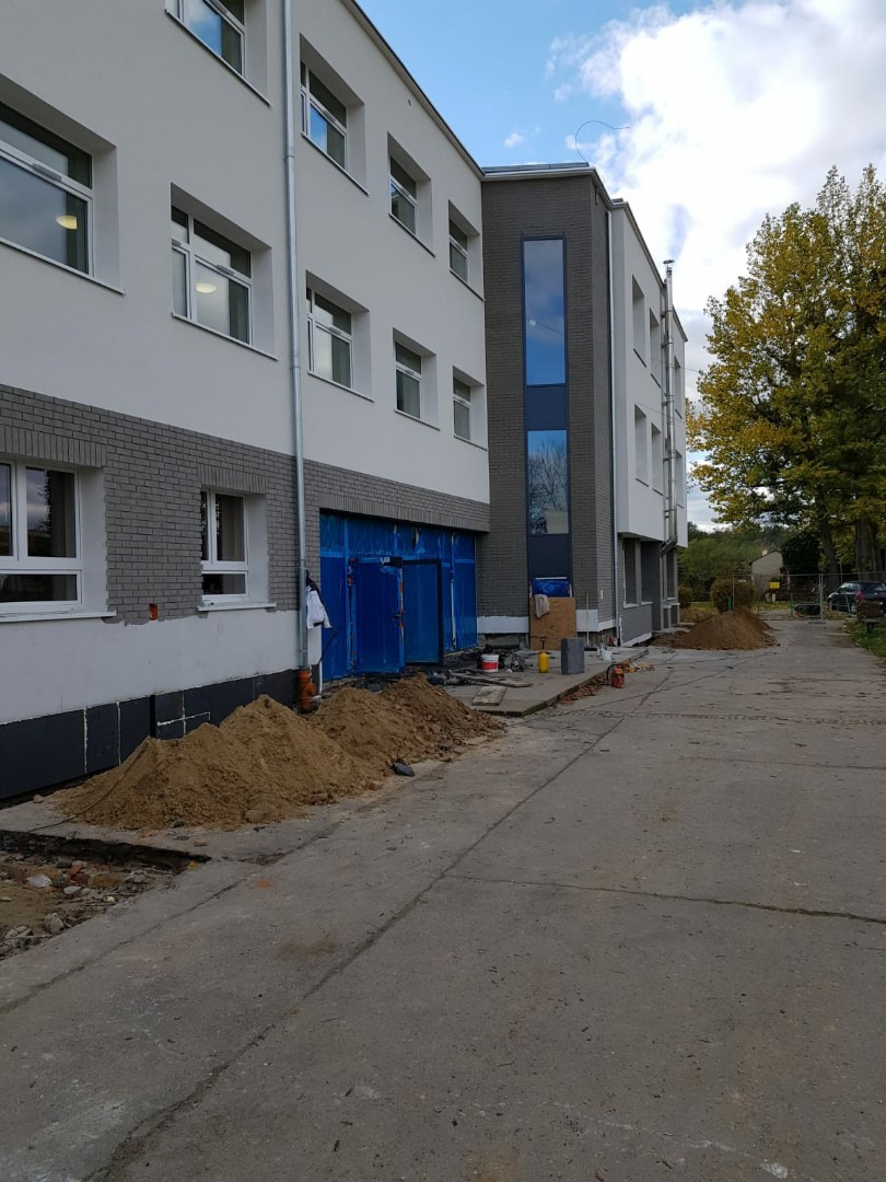 Budynek w trakcie remontu elewacji i wymiany stolarki okiennej, widoczne okna z białymi ramami, świeżo otynkowana ściana, zabezpieczone wejście i hałda piachu.