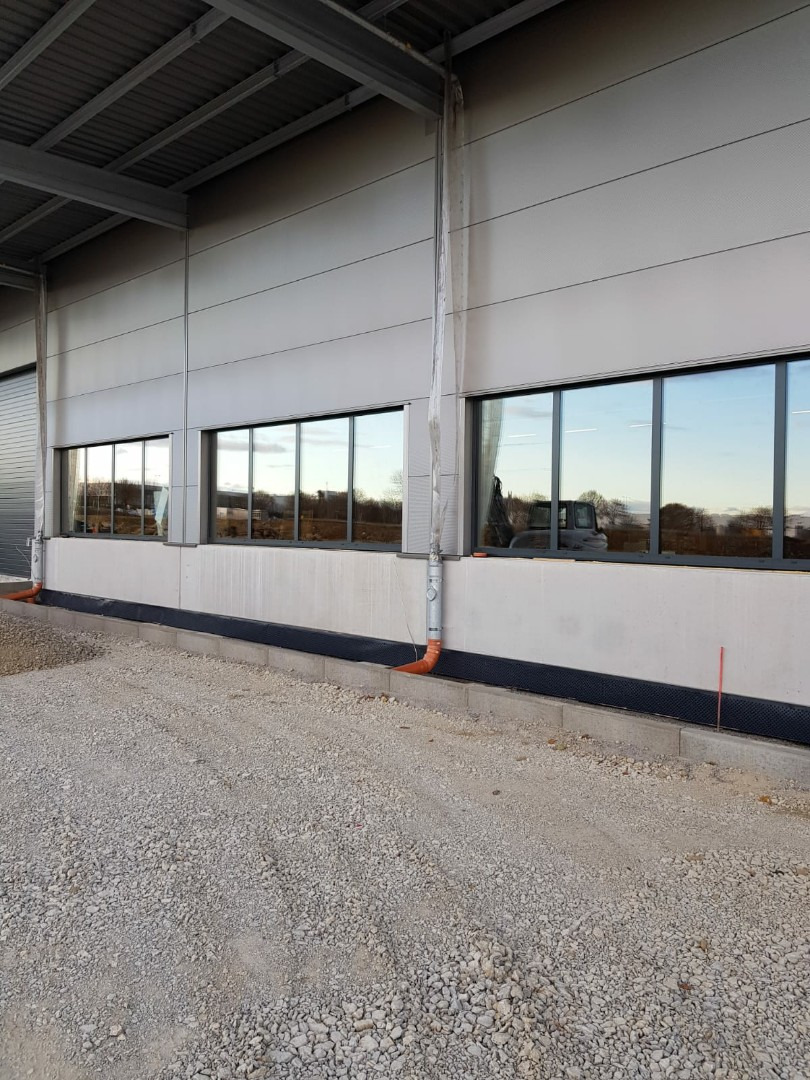 Szereg okien w metalowej elewacji budynku przemysłowego z widocznymi rynnami i żwirowym podłożem.