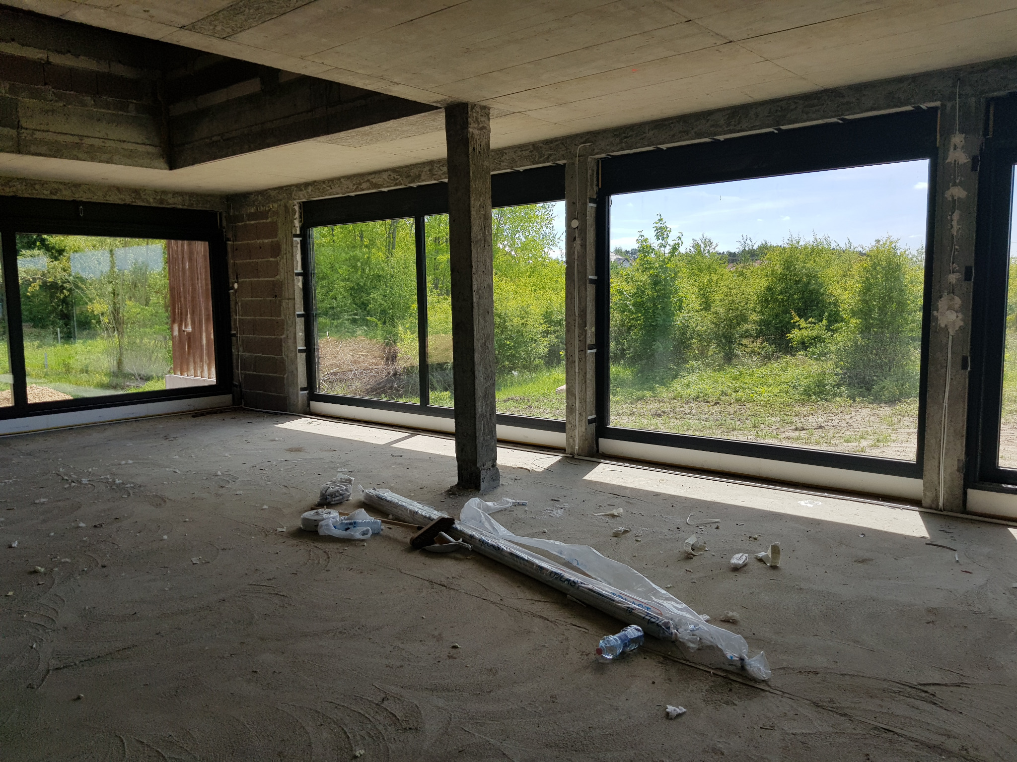 Surowy stan wykończeniowy wnętrza z dużymi oknami z czarnymi ramami, widok na zieleń za oknem, na podłodze folie i narzędzia.