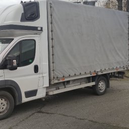 TRANS-TOM Tomasz Małka - Biały samochód dostawczy z plandeką na parkingu, widok z przodu i boku, szara plandeka, tablica rejestracyjna widoczna.