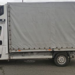 TRANS-TOM Tomasz Małka - Biały samochód dostawczy z plandeką na parkingu miejskim, widok z boku, z dodatkową przestrzenią nad kabiną.