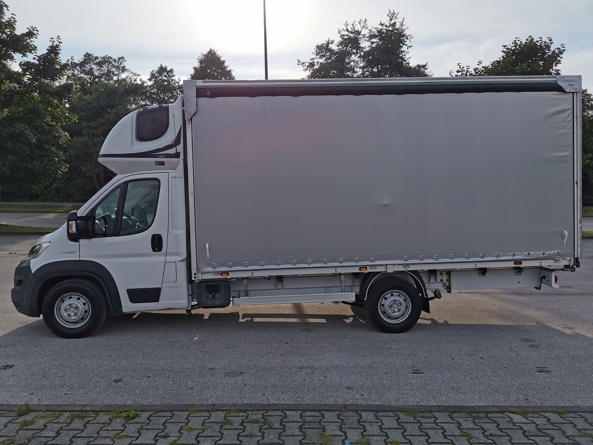 Biały samochód dostawczy z plandeką na parkingu, widok z boku, z dodatkową kabiną sypialną nad kabiną kierowcy.