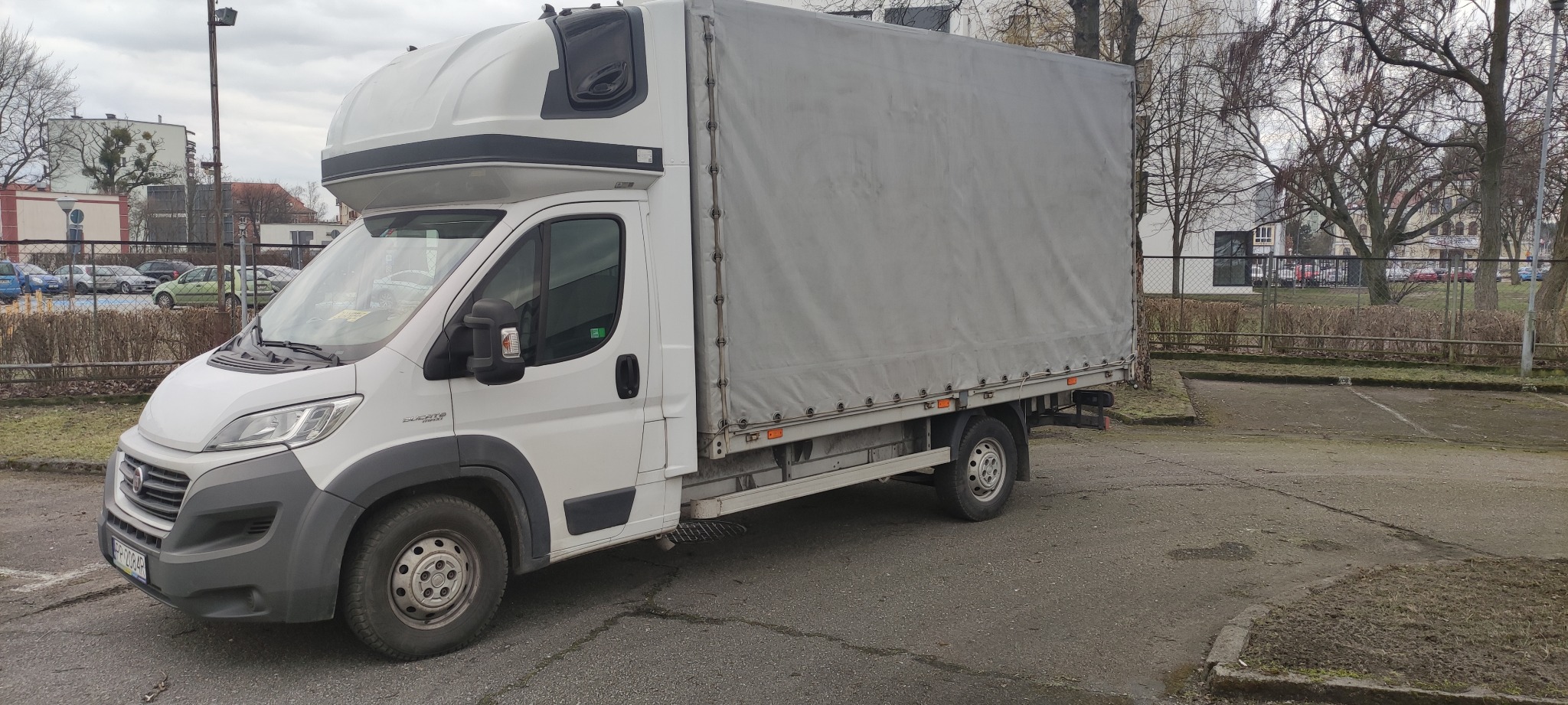 Biały samochód dostawczy z plandeką na parkingu, widok z przodu i boku, szara plandeka, tablica rejestracyjna widoczna.