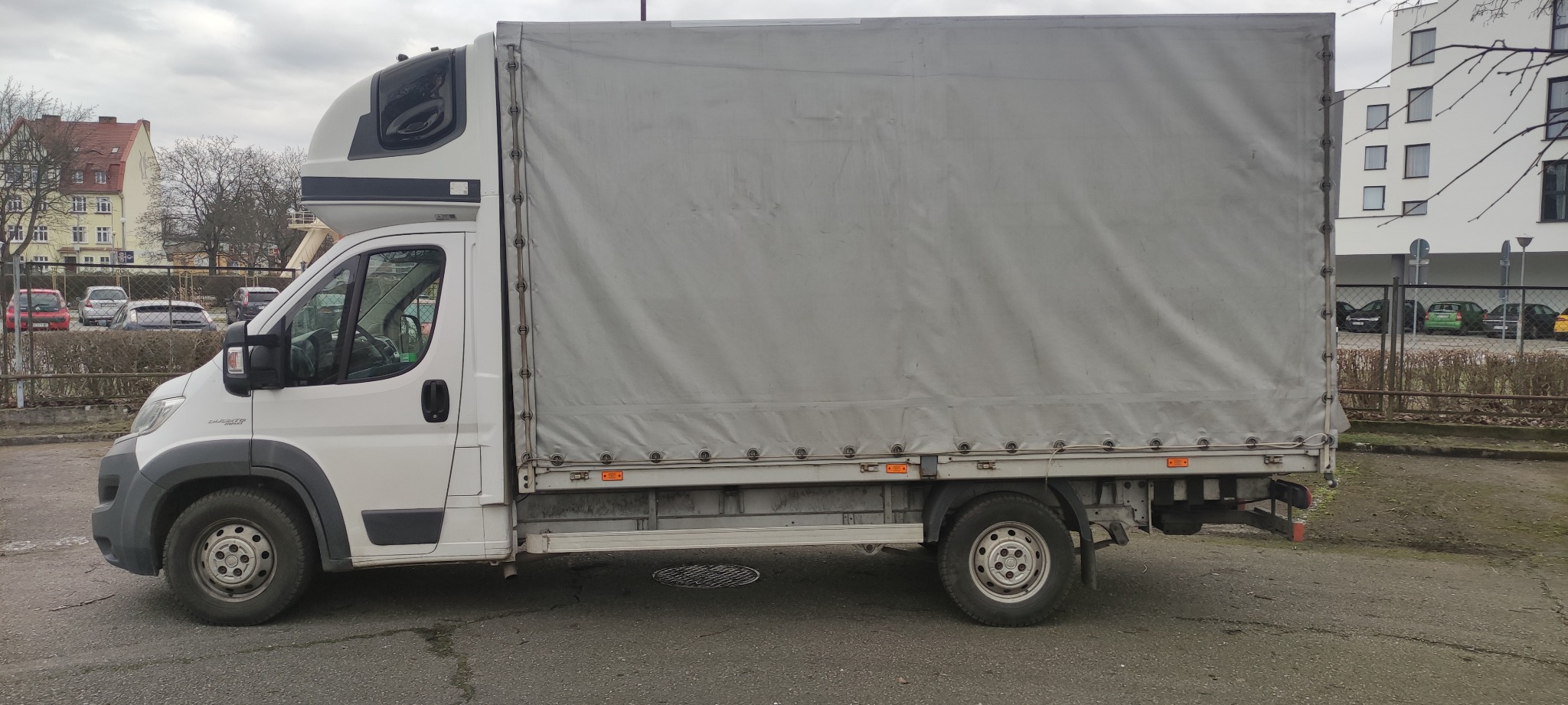 Biały samochód dostawczy z plandeką na parkingu miejskim, widok z boku, z dodatkową przestrzenią nad kabiną.