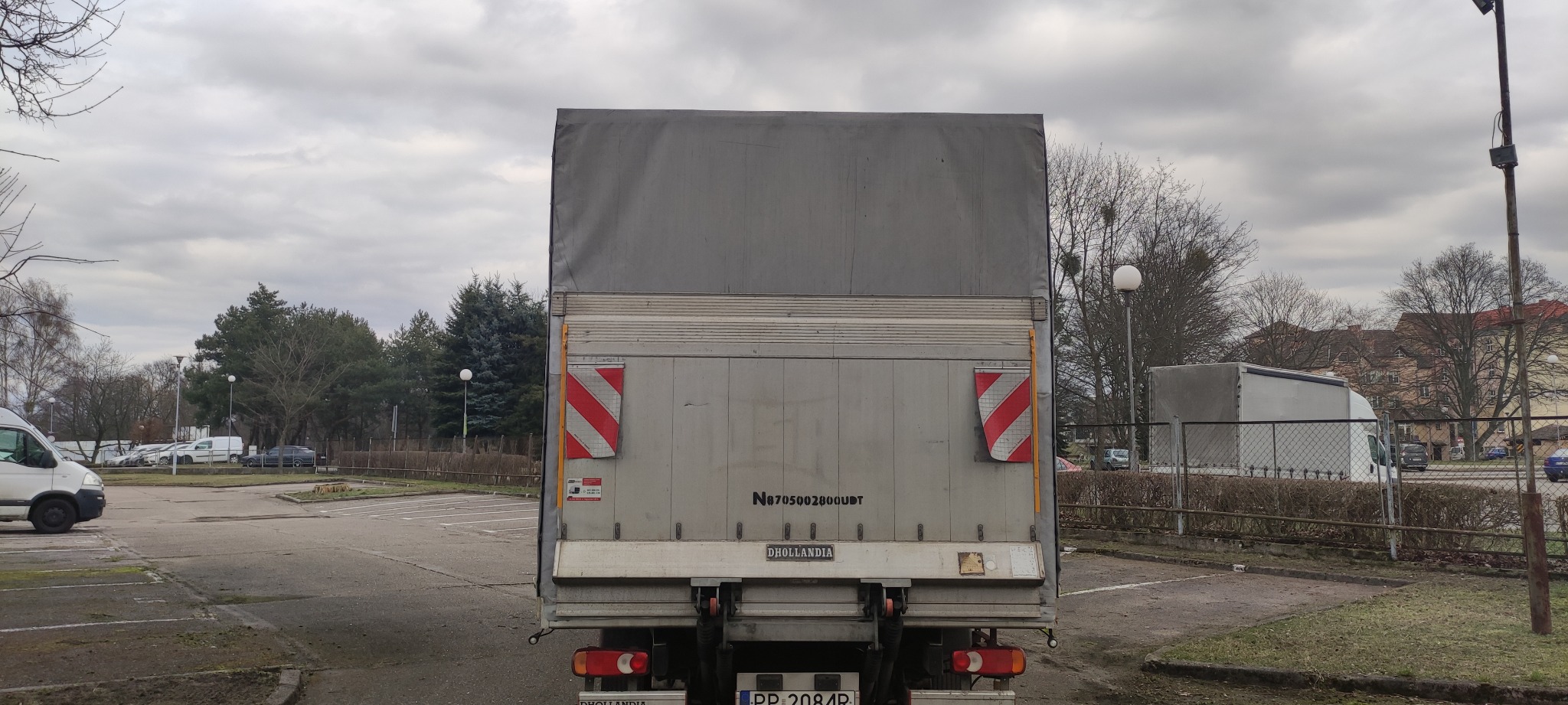 Tył szarego samochodu dostawczego z plandeką na parkingu w pochmurny dzień, widoczne tablice ostrzegawcze i tablica rejestracyjna.