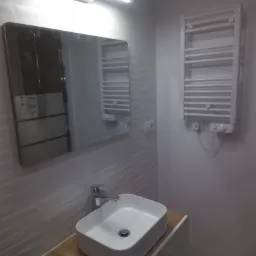 Nowoczesna łazienka z umywalką nablatową, lustrem z zaokrąglonymi rogami i grzejnikiem drabinkowym, wykończona płytkami z delikatnym wzorem.