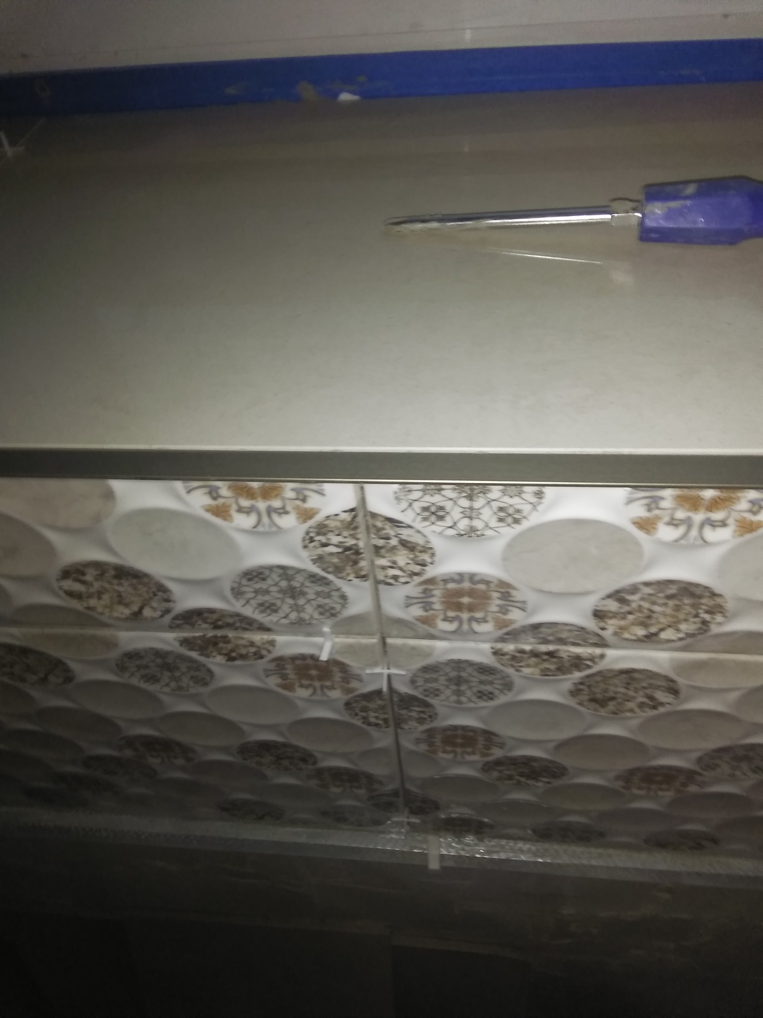 Fragment ściany z ułożonymi płytkami ceramicznymi o okrągłym wzorze, widoczny niebieski uchwyt poziomicy i szare fugi między płytkami w trakcie prac wykończeniowych.