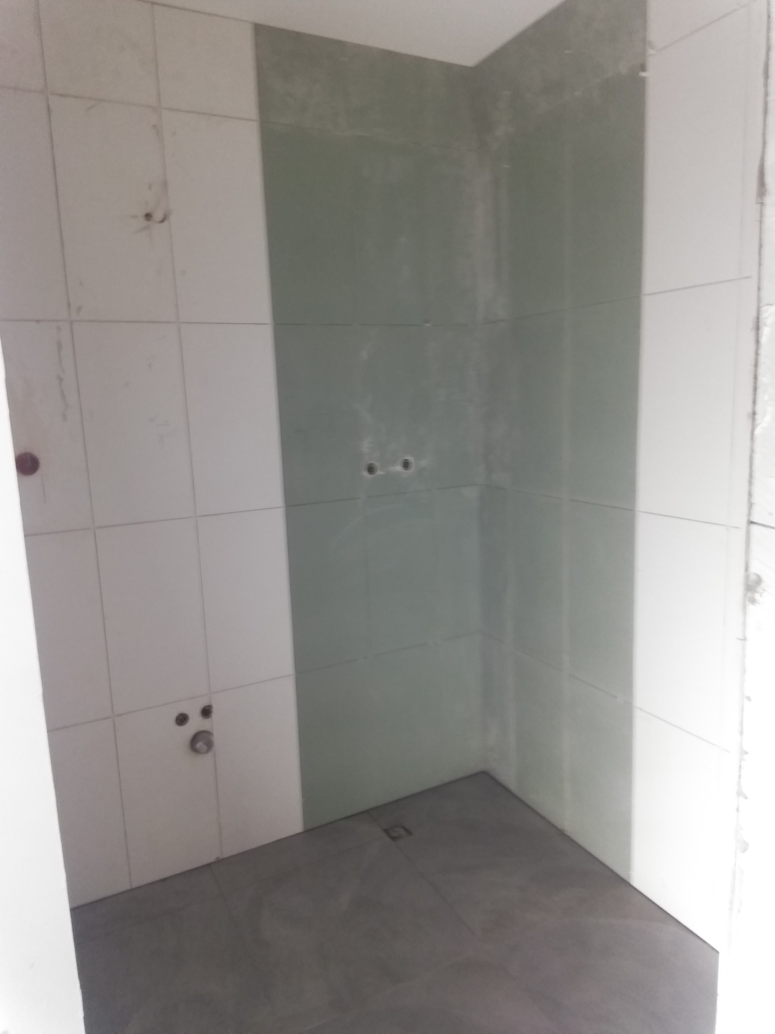 Niedokończona łazienka w trakcie remontu: białe płytki częściowo położone, szara podłoga, przygotowane ściany pod prysznic z widocznymi otworami na instalacje.