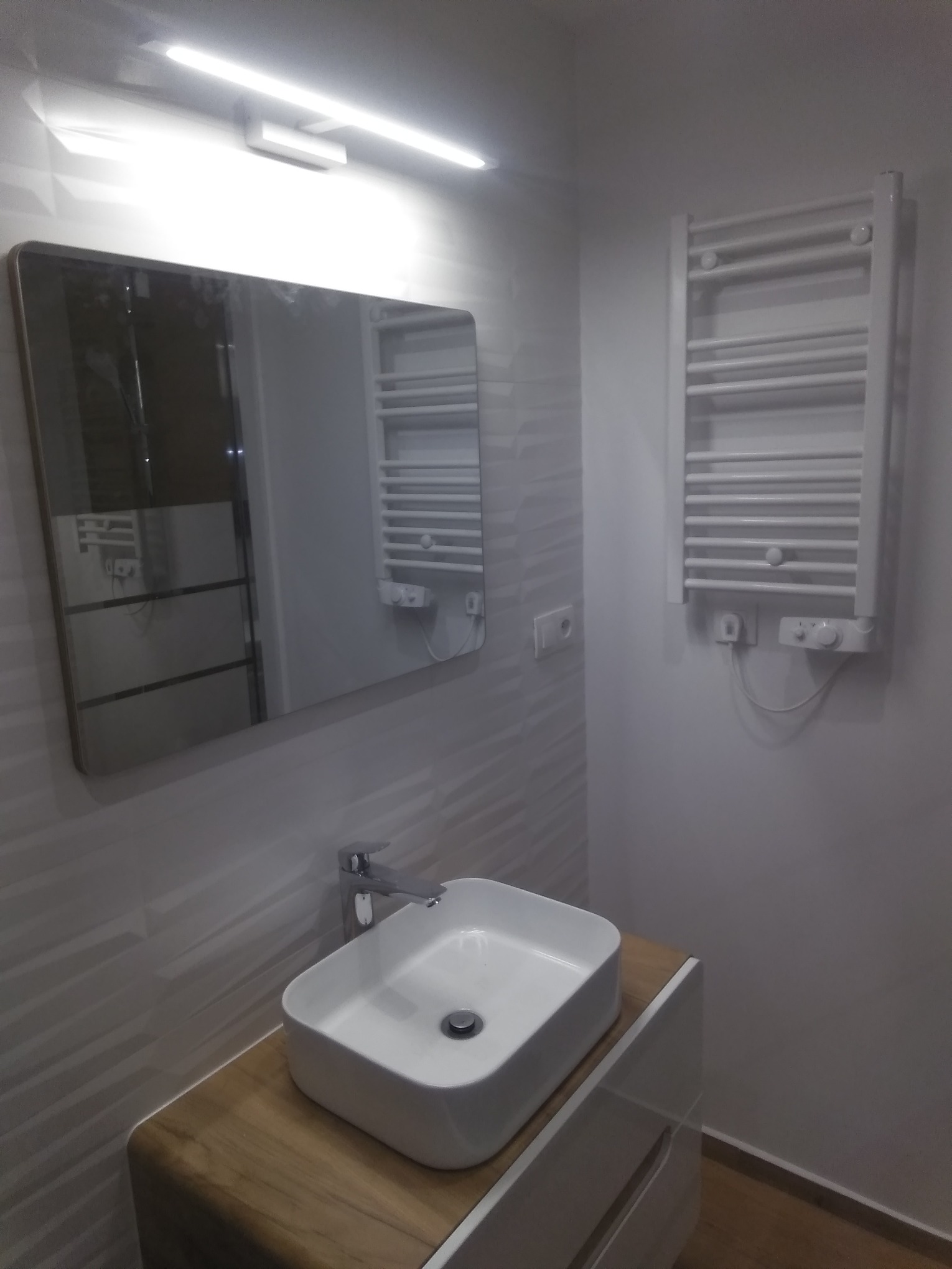 Nowoczesna łazienka z umywalką nablatową, lustrem z zaokrąglonymi rogami i grzejnikiem drabinkowym, wykończona płytkami z delikatnym wzorem.