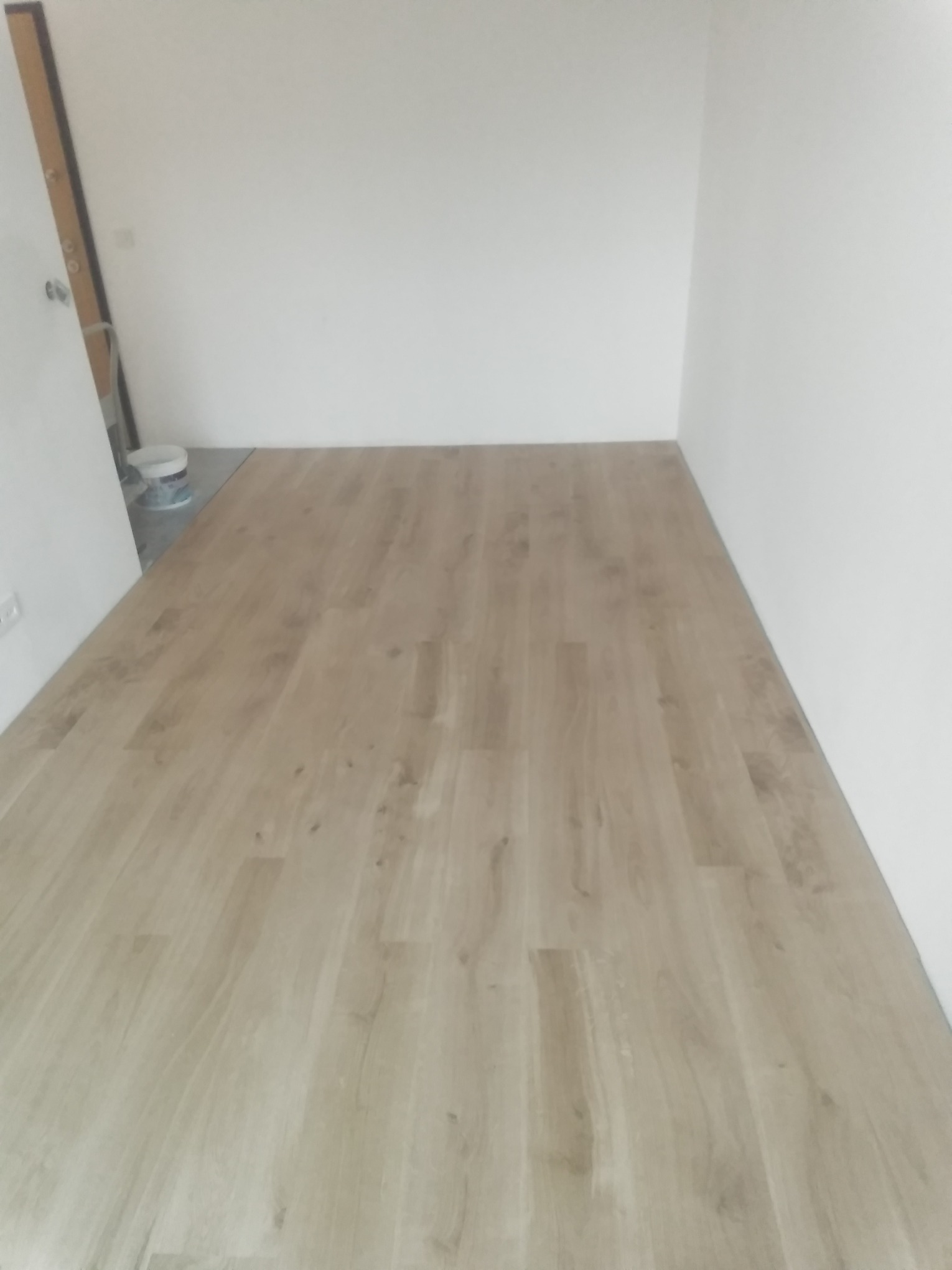 Wykończona podłoga z paneli laminowanych w jasnym odcieniu drewna, widoczny fragment białej ściany i drzwi, wiaderko z farbą w rogu pomieszczenia.
