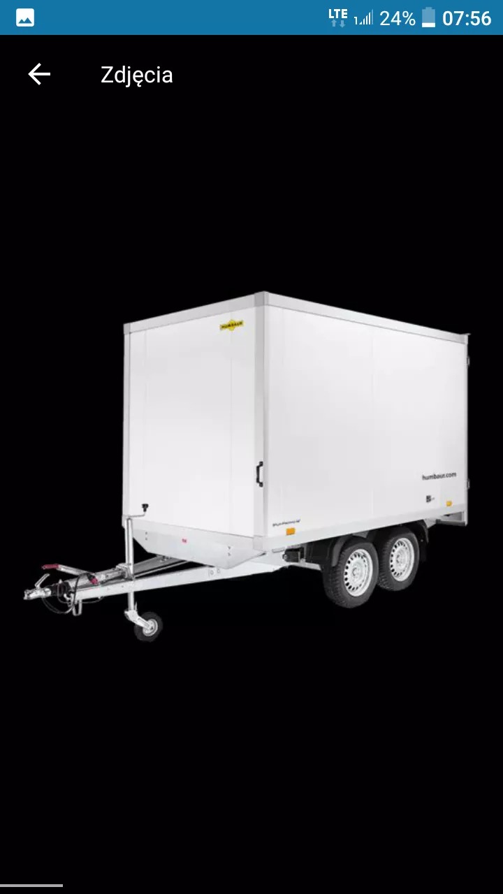 Biała, zamknięta przyczepka kontenerowa z dwoma osiami, widoczny zaczep i opony z bieżnikiem, na czarnym tle.