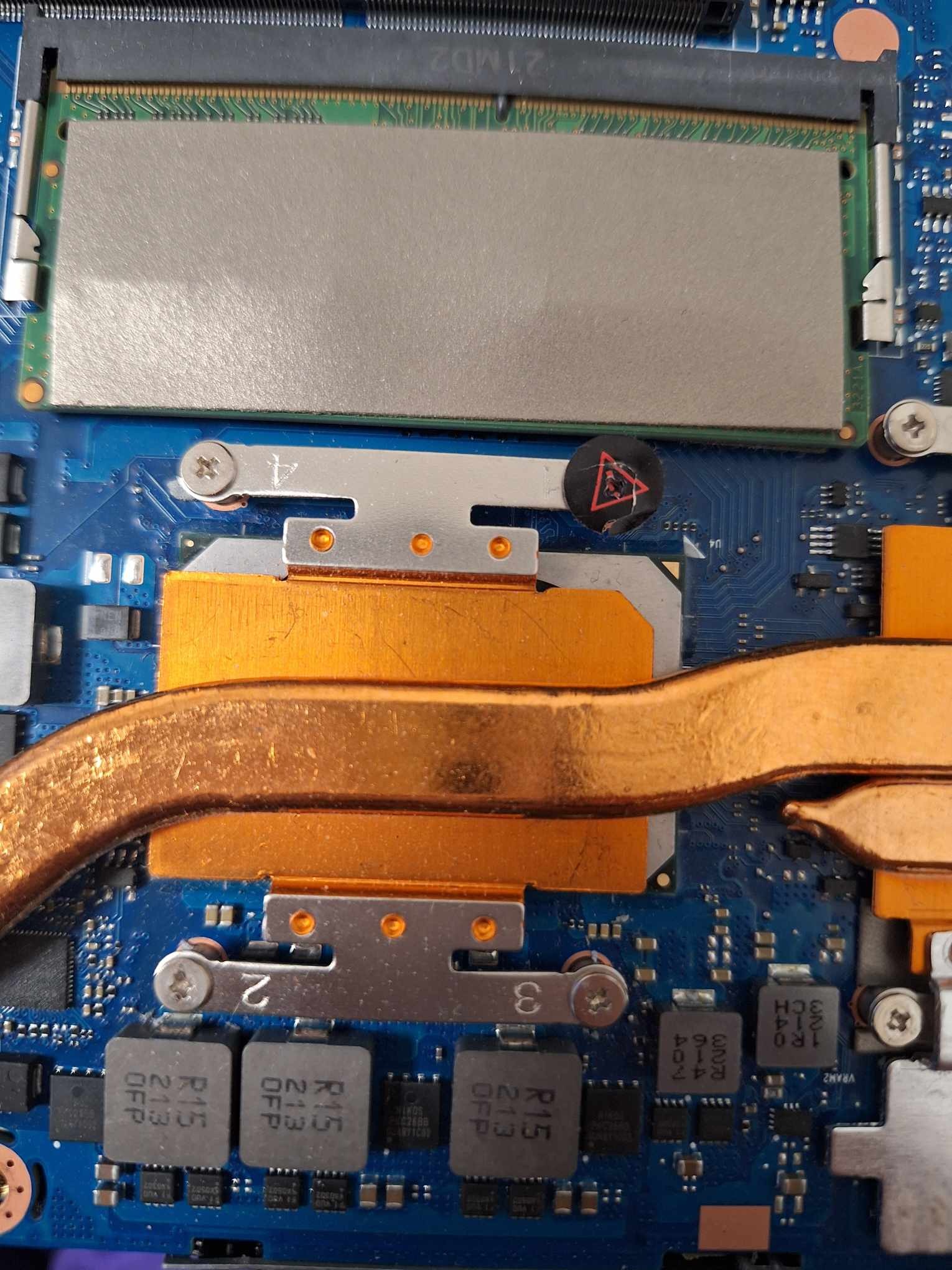 Wnętrze laptopa z widocznym układem chłodzenia (miedziane rurki cieplne i radiatory) oraz kością pamięci RAM, niebieska płyta główna z elementami elektronicznymi i naklejką ostrzegawczą z symbolem...