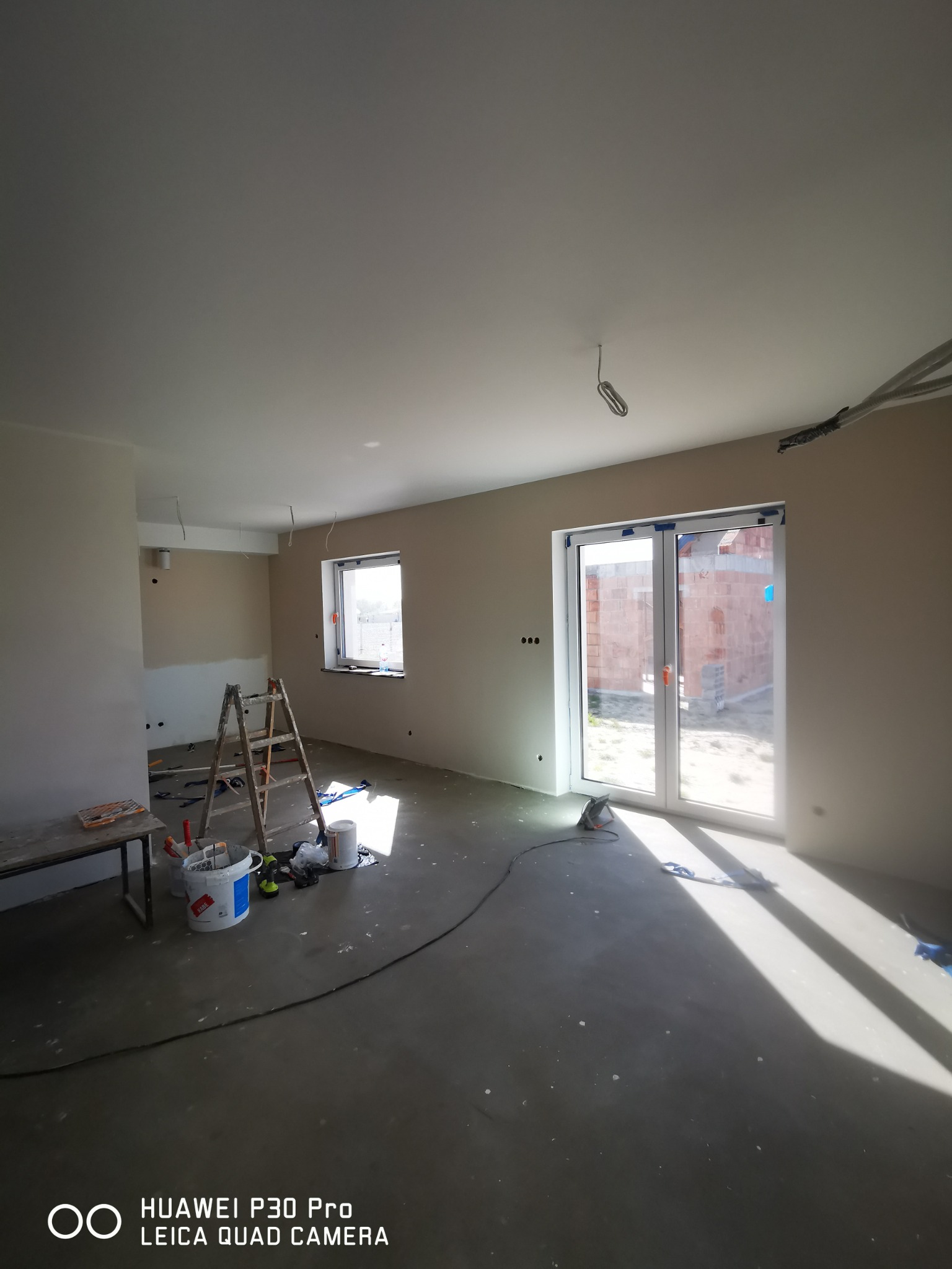 Wnętrze w trakcie malowania: widoczne ściany po szpachlowaniu, drabina, pędzle, wiaderka z farbą oraz okno z widokiem na budynek w trakcie budowy.