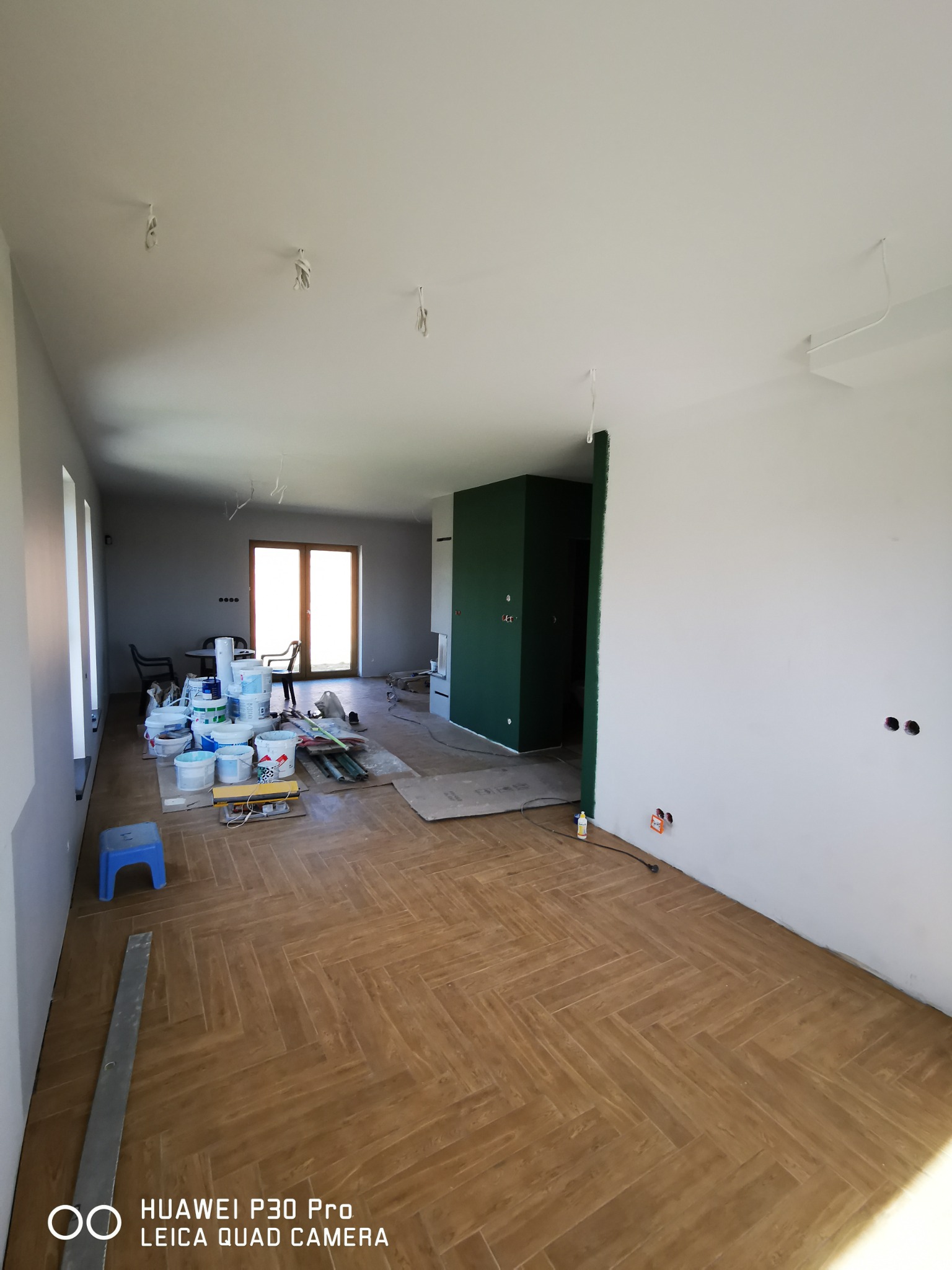 Wnętrze w trakcie malowania: widoczne wiadra z farbą, narzędzia malarskie i fragment pomalowanej na zielono ściany w sąsiednim pomieszczeniu; na suficie zwisające przewody oświetleniowe.