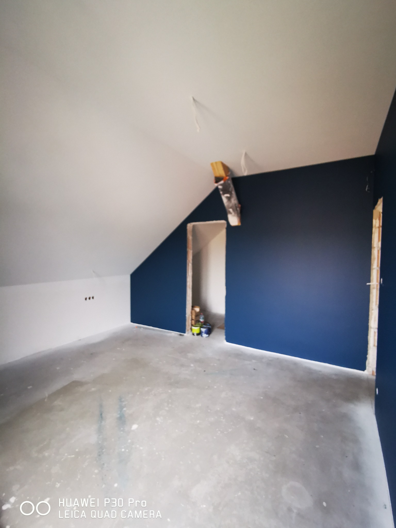 Pomieszczenie na poddaszu w trakcie malowania we Wrześni: ściany częściowo pomalowane na granatowo, sufit biały, widoczne drabiny i puszki z farbą.