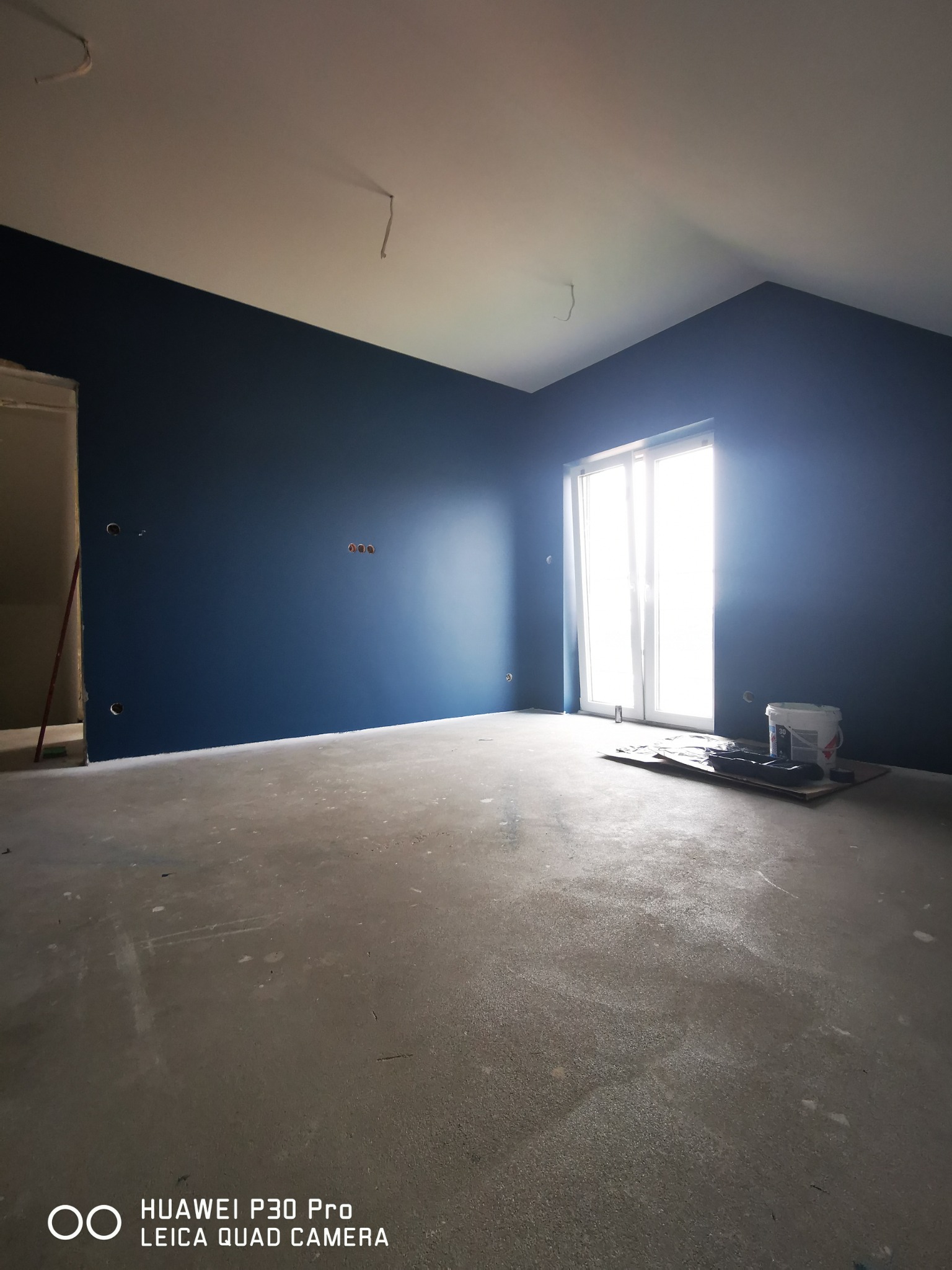 Pomieszczenie po malowaniu ścian na ciemny niebieski kolor, widoczne puszki z farbą przy oknie, surowa podłoga betonowa, przygotowane otwory na gniazdka elektryczne.