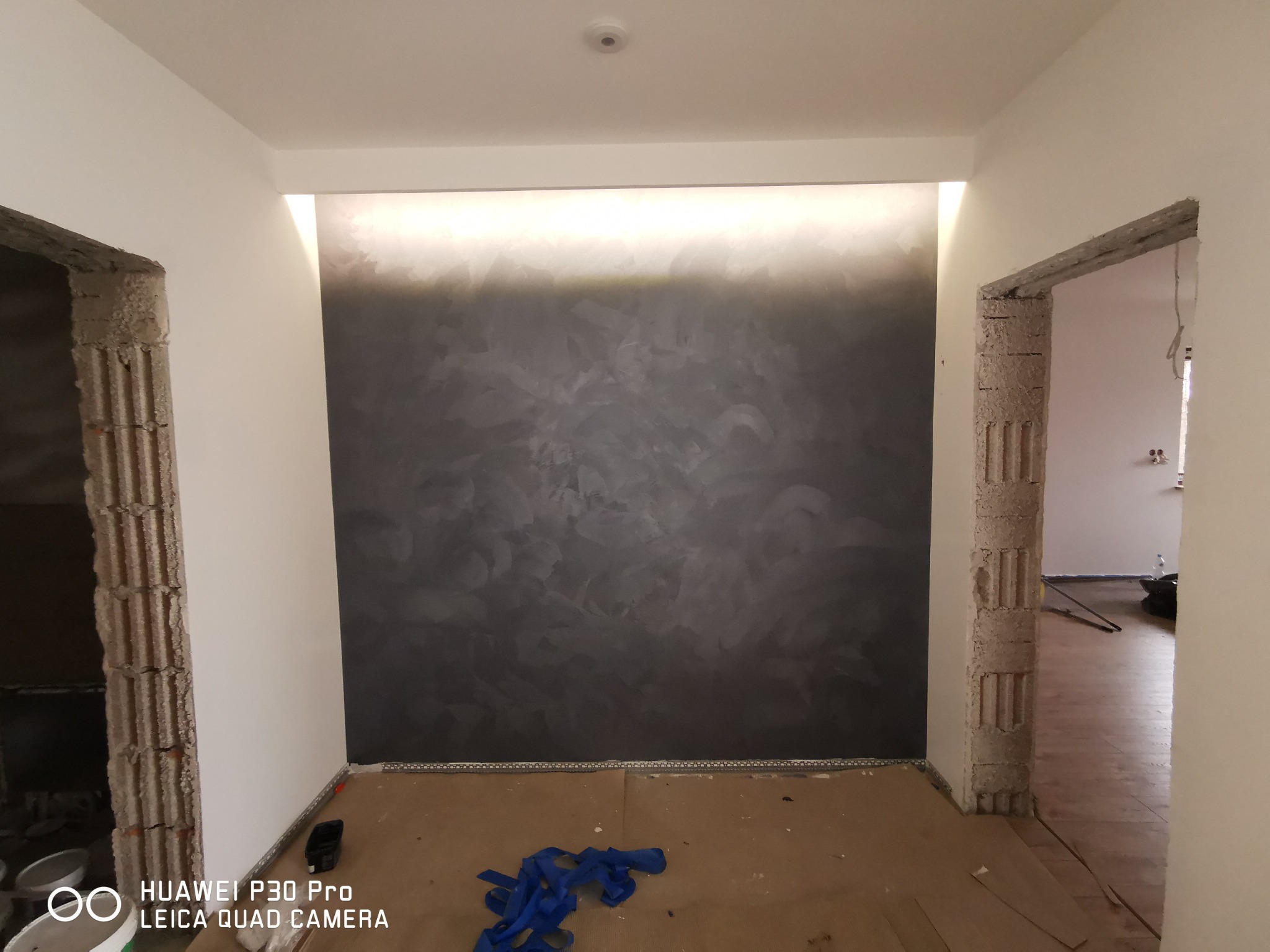 Pokój w trakcie remontu: ściana z dekoracyjnym szarym wykończeniem, oświetlenie LED podsufitowe, surowe ościeżnice drzwiowe i zabezpieczona podłoga.