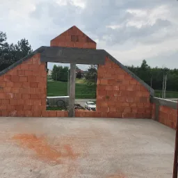 Surowy stan poddasza z czerwonej cegły ceramicznej, widok przez otwór okienny na krajobraz z białym samochodem, fragment rusztowania po prawej.