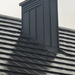 Szary, metalowy komin wentylacyjny na dachu pokrytym ciemną dachówką ceramiczną.