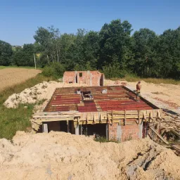 Budowa domu jednorodzinnego: wzniesione ściany fundamentowe z czerwonej cegły, szalunek na stropie oraz fragment wzniesionej ściany parteru w tle, na działce z widokiem na pole i las.