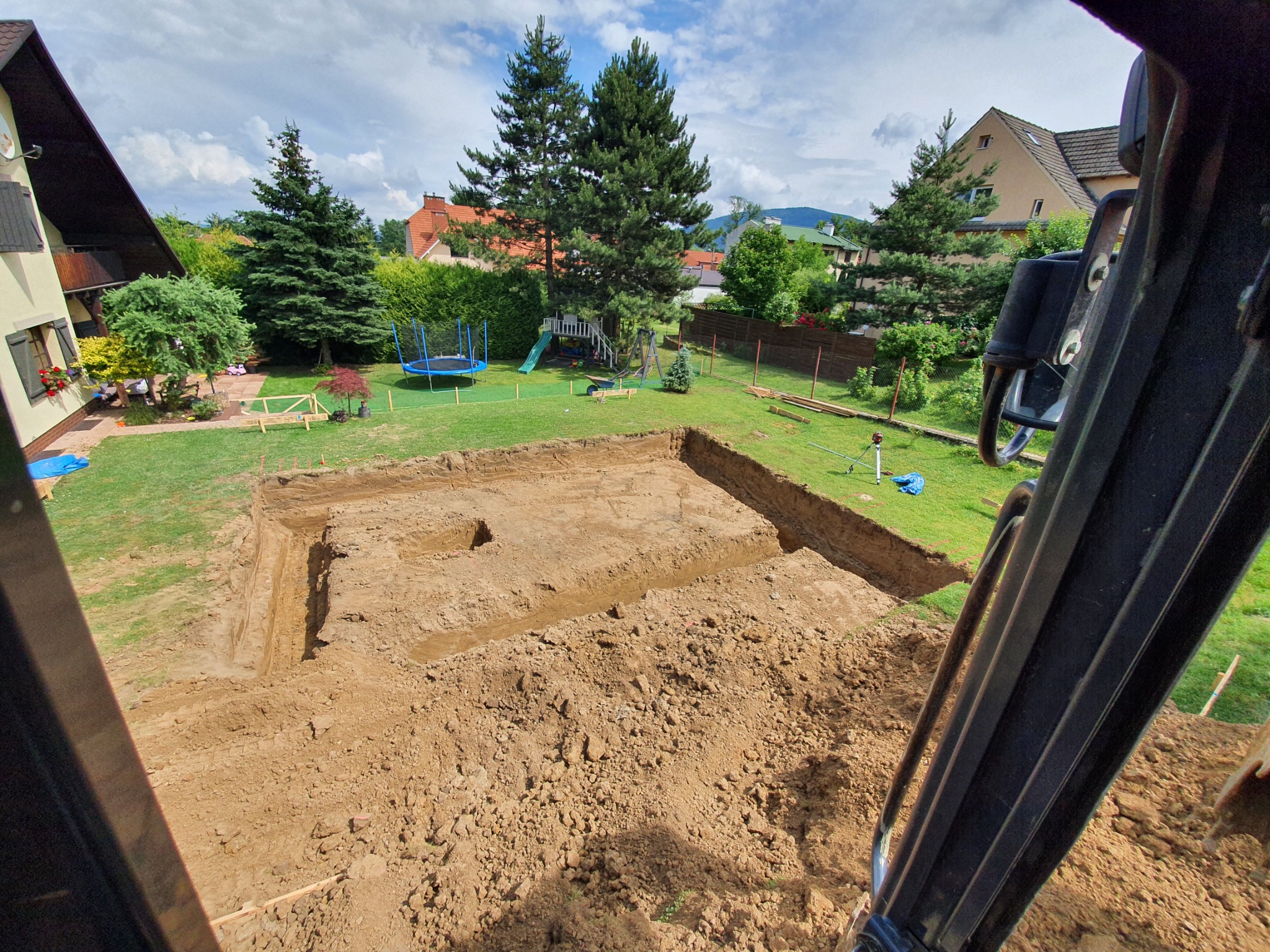 Wykopany fundament pod budynek jednorodzinny, widok z perspektywy kabiny koparki, w tle trawnik z trampoliną i drzewami.