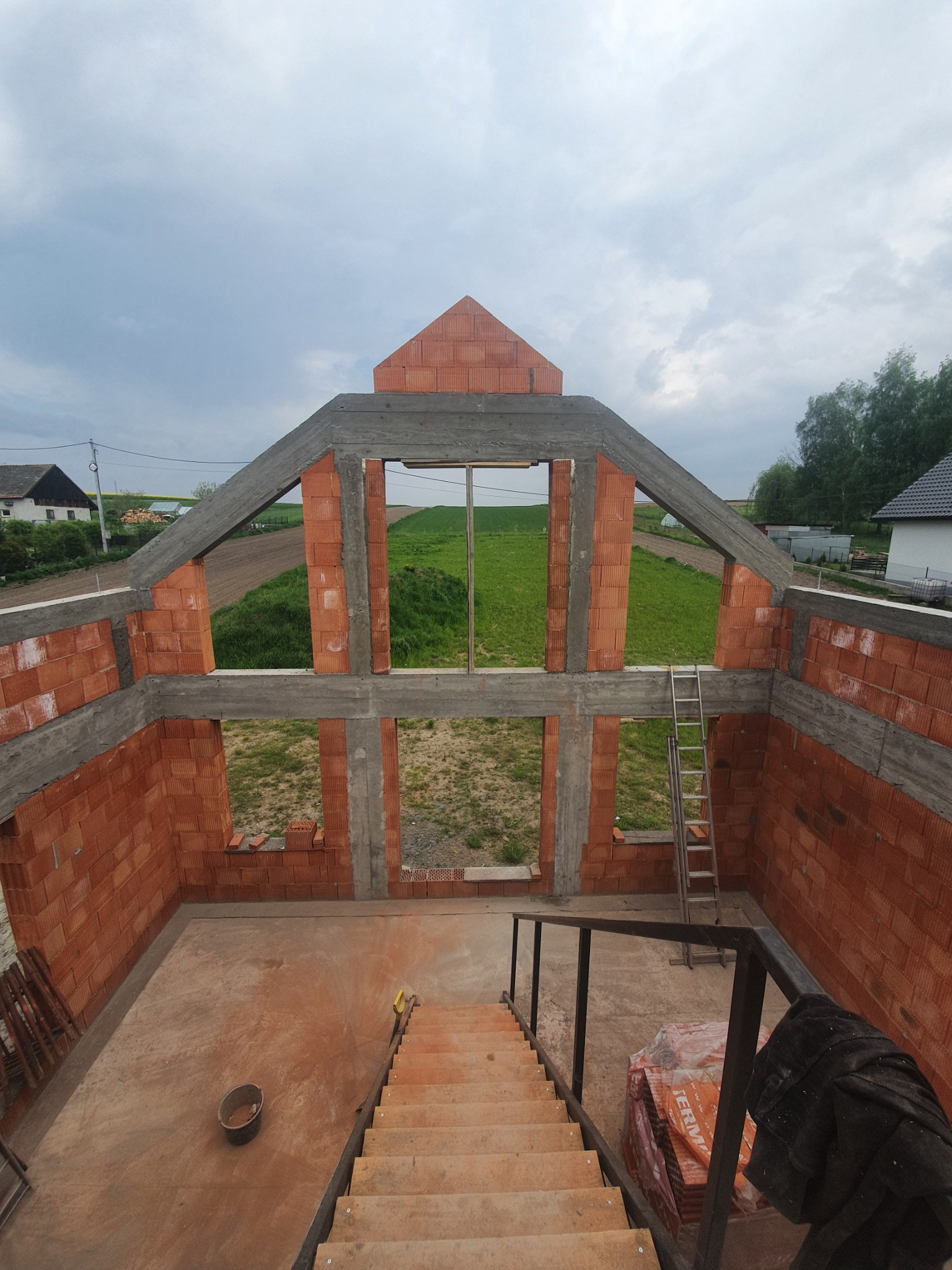 Wnętrze murowanego domu w trakcie budowy, widoczne ściany z czerwonej cegły, betonowe nadproża, schody tymczasowe i drabina oparta o ścianę, w tle zielone pola i niebo z chmurami.