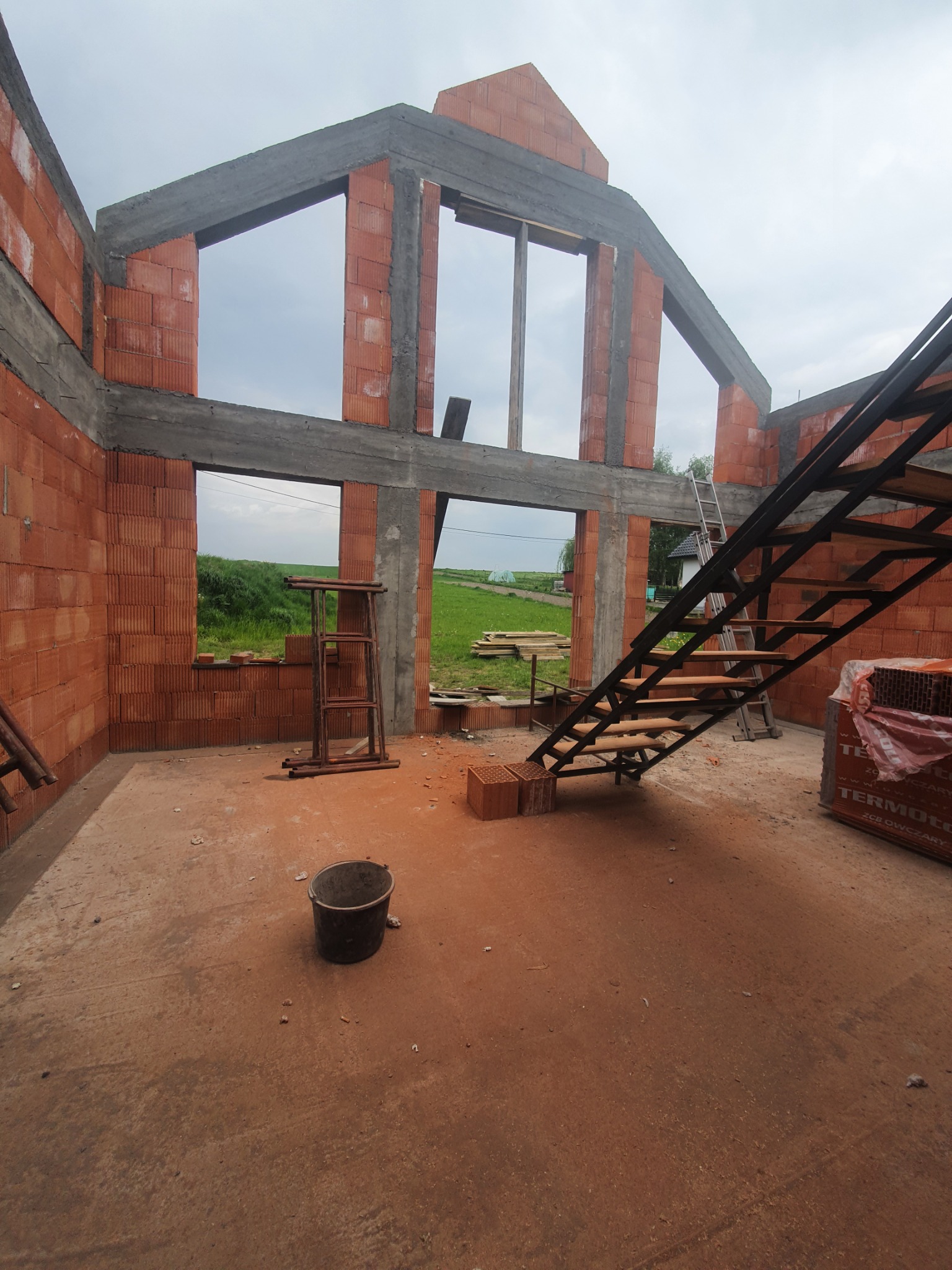 Wnętrze domu w trakcie budowy z czerwonej cegły, widoczne schody stalowe, drabina oparta o ścianę, wiadro i paleta z cegłami w tle, otwarty krajobraz za oknami.