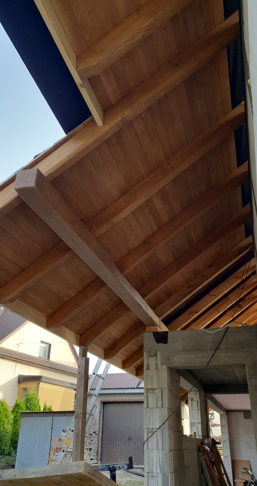 Drewniany strop w trakcie budowy, widoczne belki, deski, fragment ściany z betonu komórkowego oraz drabina oparta o ścianę budynku w tle.