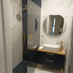 Nowoczesna łazienka z kabiną prysznicową typu walk-in, biało-marmurkowymi płytkami ściennymi, granatowymi płytkami ułożonymi w jodełkę pod prysznicem, okrągłym lustrem z odbijającą się osobą...