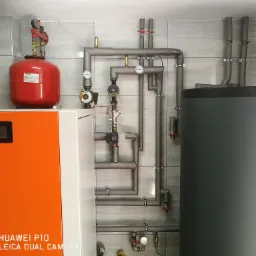 Kompletna instalacja centralnego ogrzewania z piecem, zbiornikiem buforowym Kospel, czerwonym naczyniem przeponowym Ferro i izolowanymi rurami na tle szarej ściany z płytek.