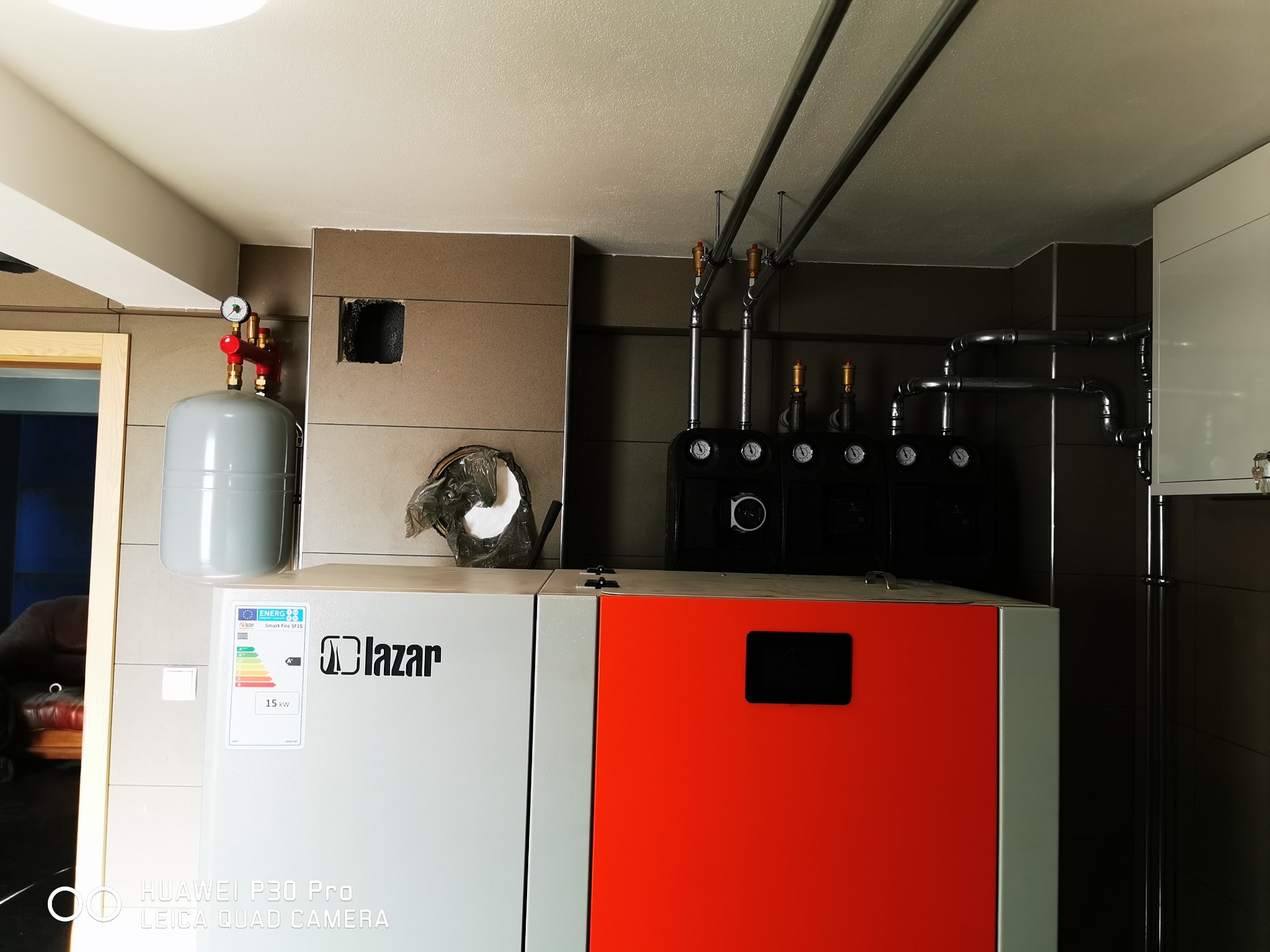 Nowoczesny piec z czerwonym panelem, szary zbiornik wyrównawczy z manometrem, instalacja rurowa pod sufitem i brązowe płytki na ścianach w kotłowni.