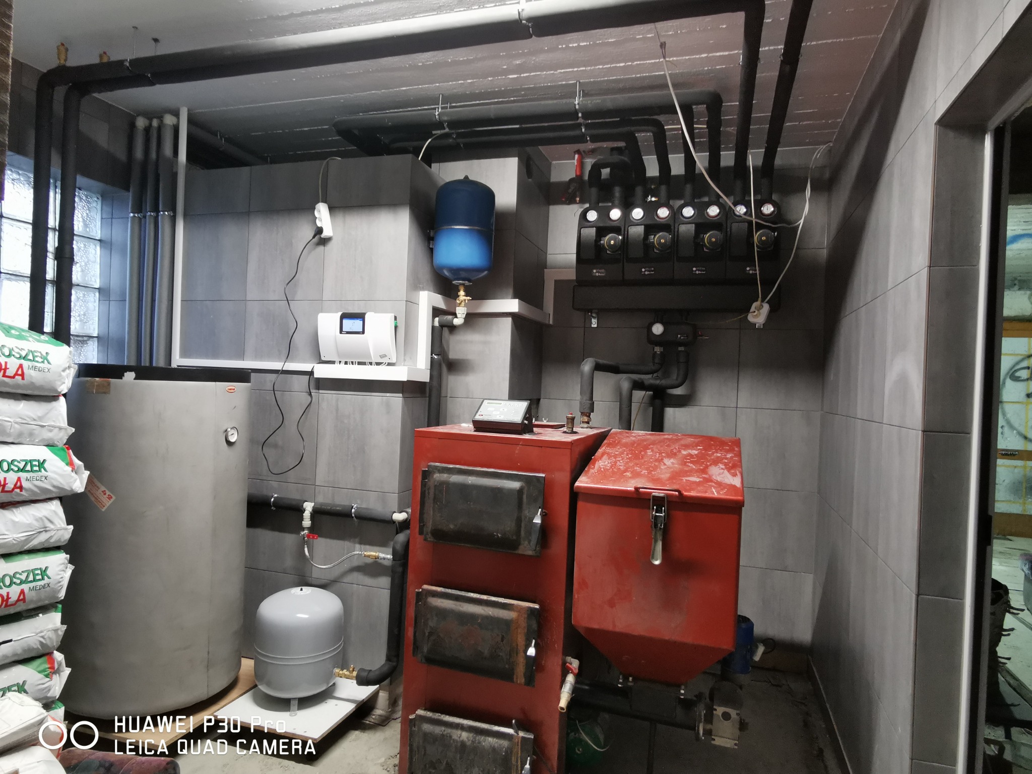 Kotłownia z kotłem na paliwo stałe, zasobnikiem ciepłej wody, zbiornikiem wyrównawczym i rozdzielaczem, widoczne worki z ekogroszkiem.