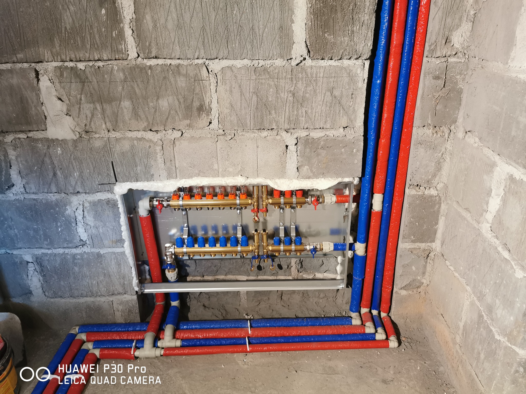 Rozdzielacz ogrzewania podłogowego z rurami w kolorach czerwonym i niebieskim, zamontowany na surowej ścianie z betonu komórkowego, widoczne zawory i przyłącza.