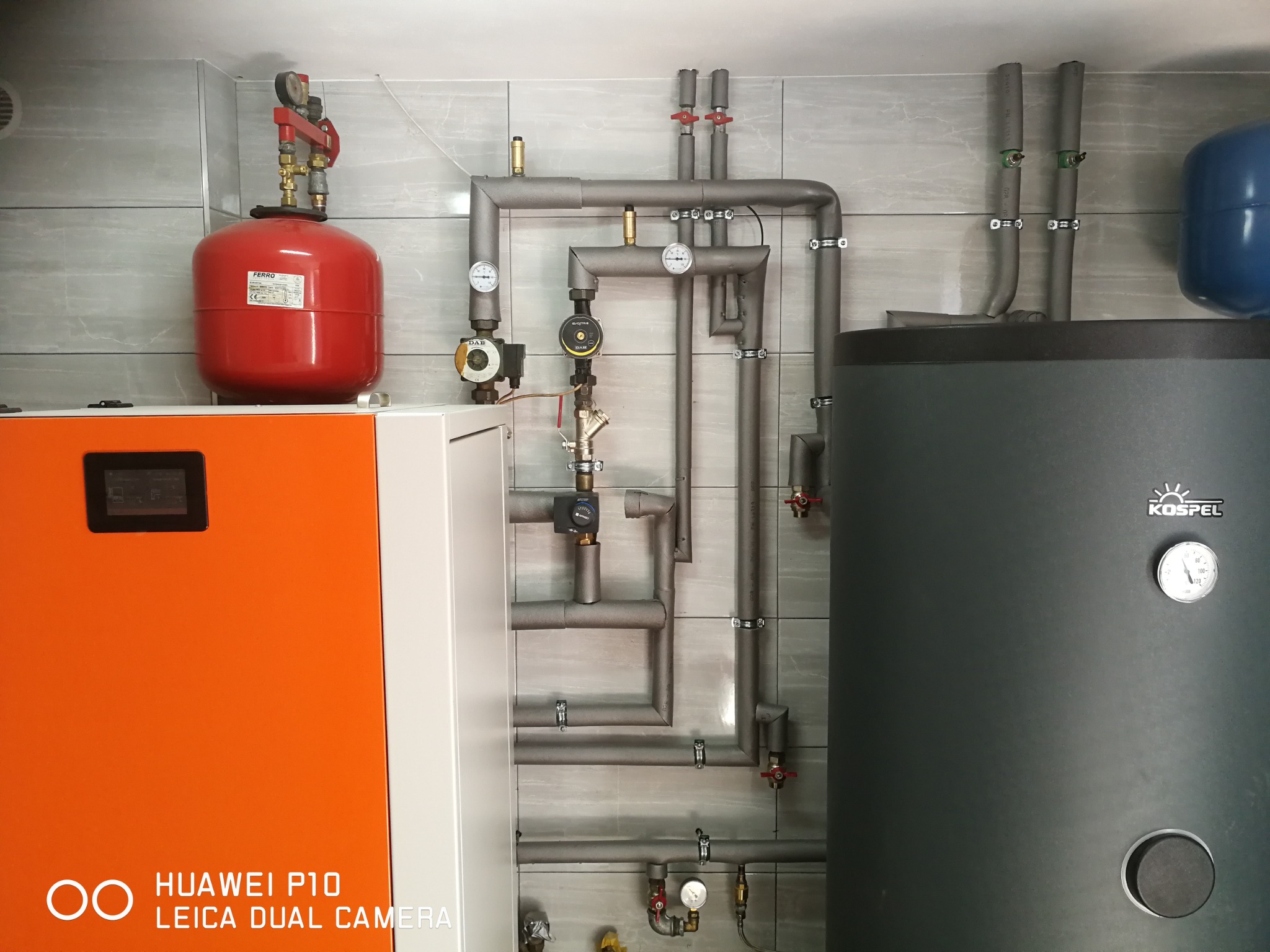 Kompletna instalacja centralnego ogrzewania z piecem, zbiornikiem buforowym Kospel, czerwonym naczyniem przeponowym Ferro i izolowanymi rurami na tle szarej ściany z płytek.
