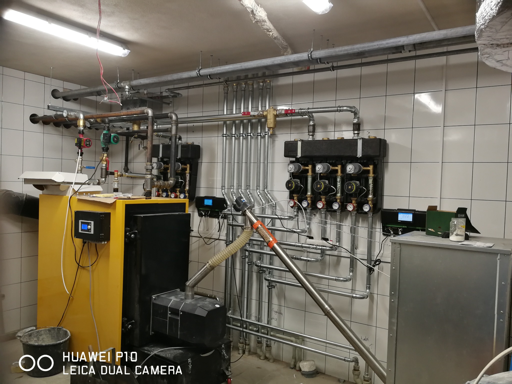 Żółty piec na paliwo stałe z automatycznym podajnikiem, widoczne rury instalacji grzewczej i zawory w kotłowni wyłożonej białymi kafelkami.