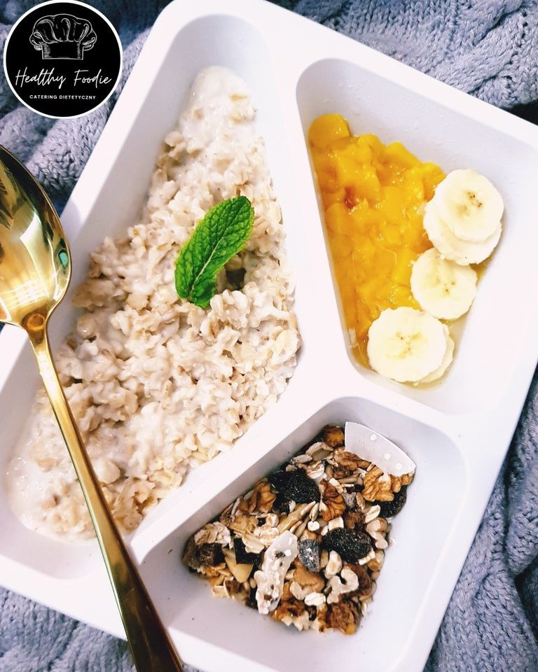 Posiłek w białym, trójdzielnym pojemniku: owsianka z miętą, mango z bananami oraz mieszanka orzechów i suszonych owoców; złota łyżka leży obok, całość na szarym kocu.