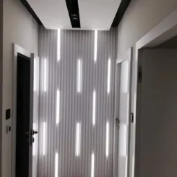 Nowoczesne wykończenie wnętrza: białe panele ścienne z pionowymi paskami LED, czarne listwy sufitowe, widoczne drzwi i fragment podłogi zabezpieczonej folią.