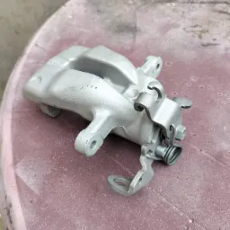 Szary, metalowy zacisk hamulcowy po renowacji, leżący na brudnym, różowym stole z tworzywa sztucznego. Widoczne ślady wcześniejszego użytkowania na zacisku.