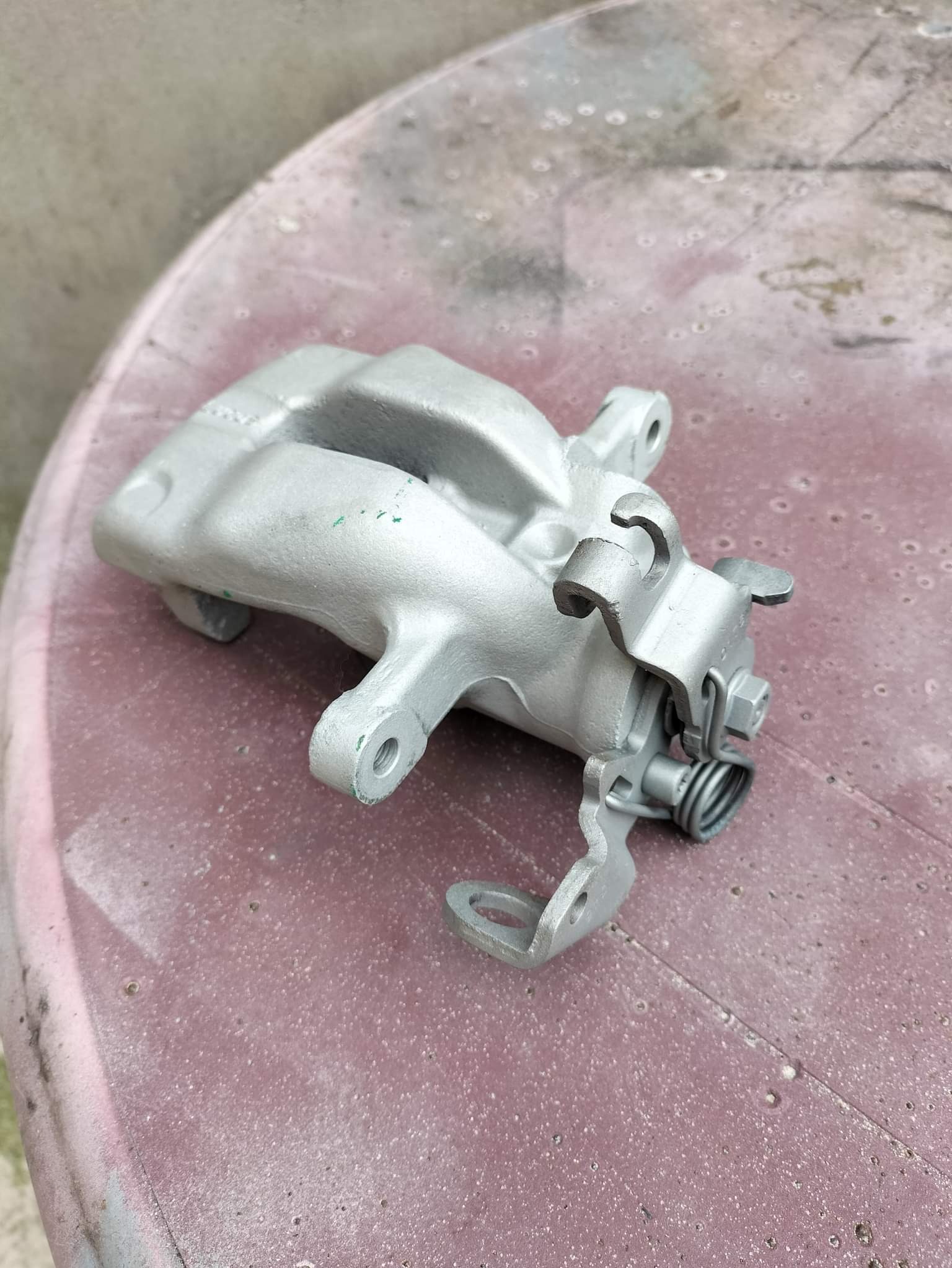Szary, metalowy zacisk hamulcowy po renowacji, leżący na brudnym, różowym stole z tworzywa sztucznego. Widoczne ślady wcześniejszego użytkowania na zacisku.