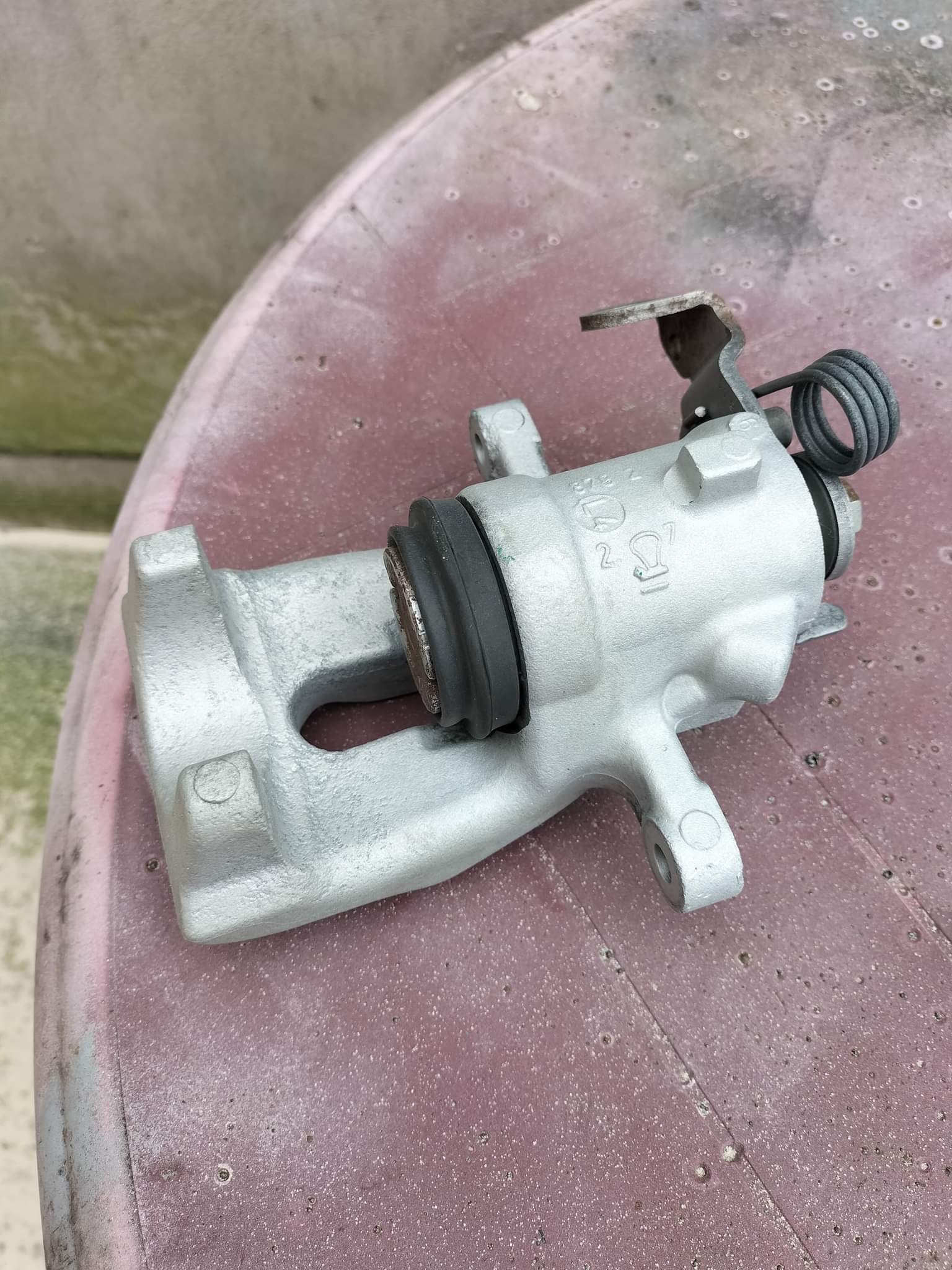 Szary zacisk hamulcowy z widocznymi oznaczeniami producenta i sprężyną, leżący na czerwonym, chropowatym stole.