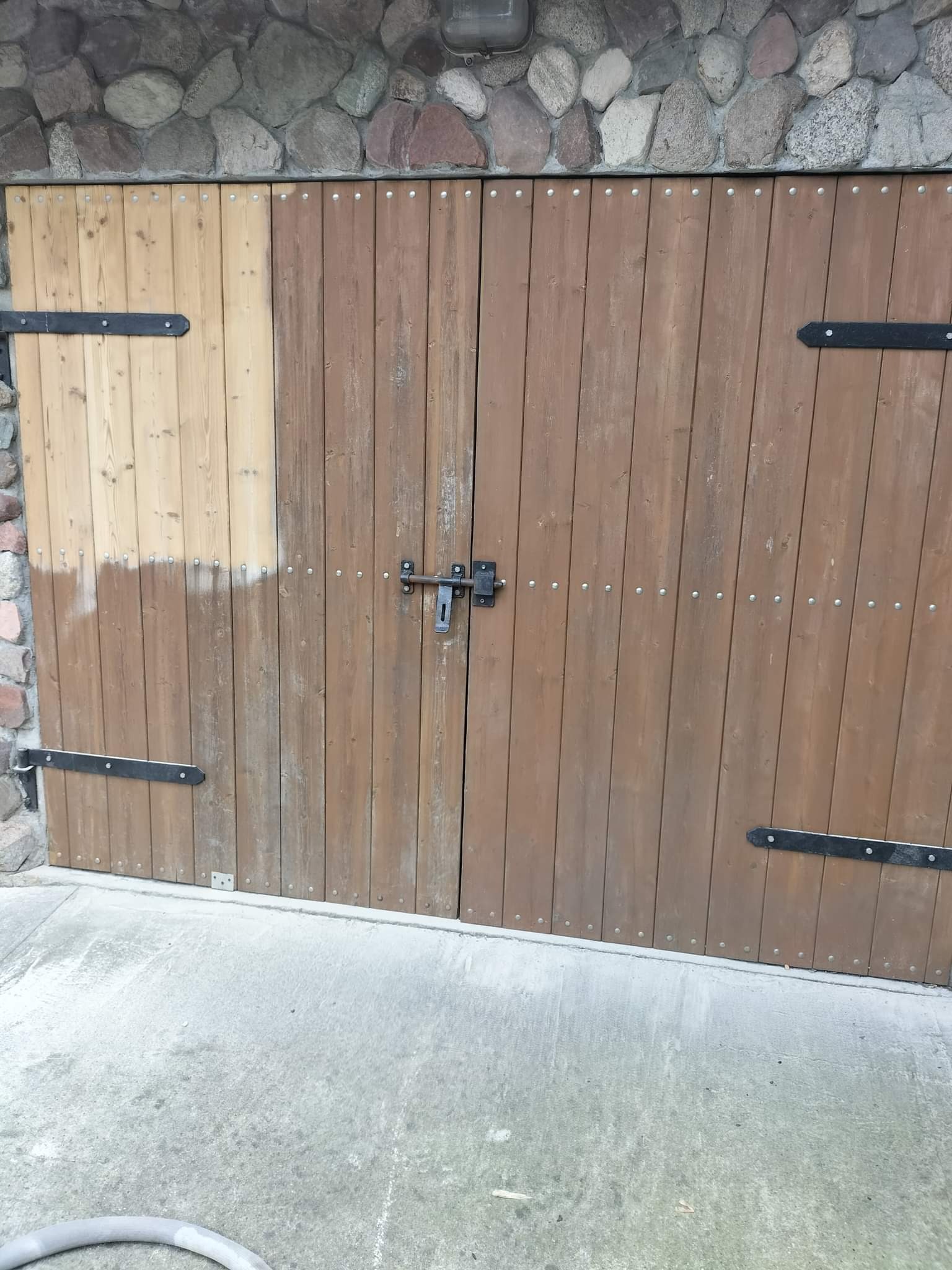 Dwuskrzydłowe drewniane drzwi z metalowymi okuciami, częściowo odnowione, z widocznym fragmentem surowego drewna po usunięciu starej powłoki.