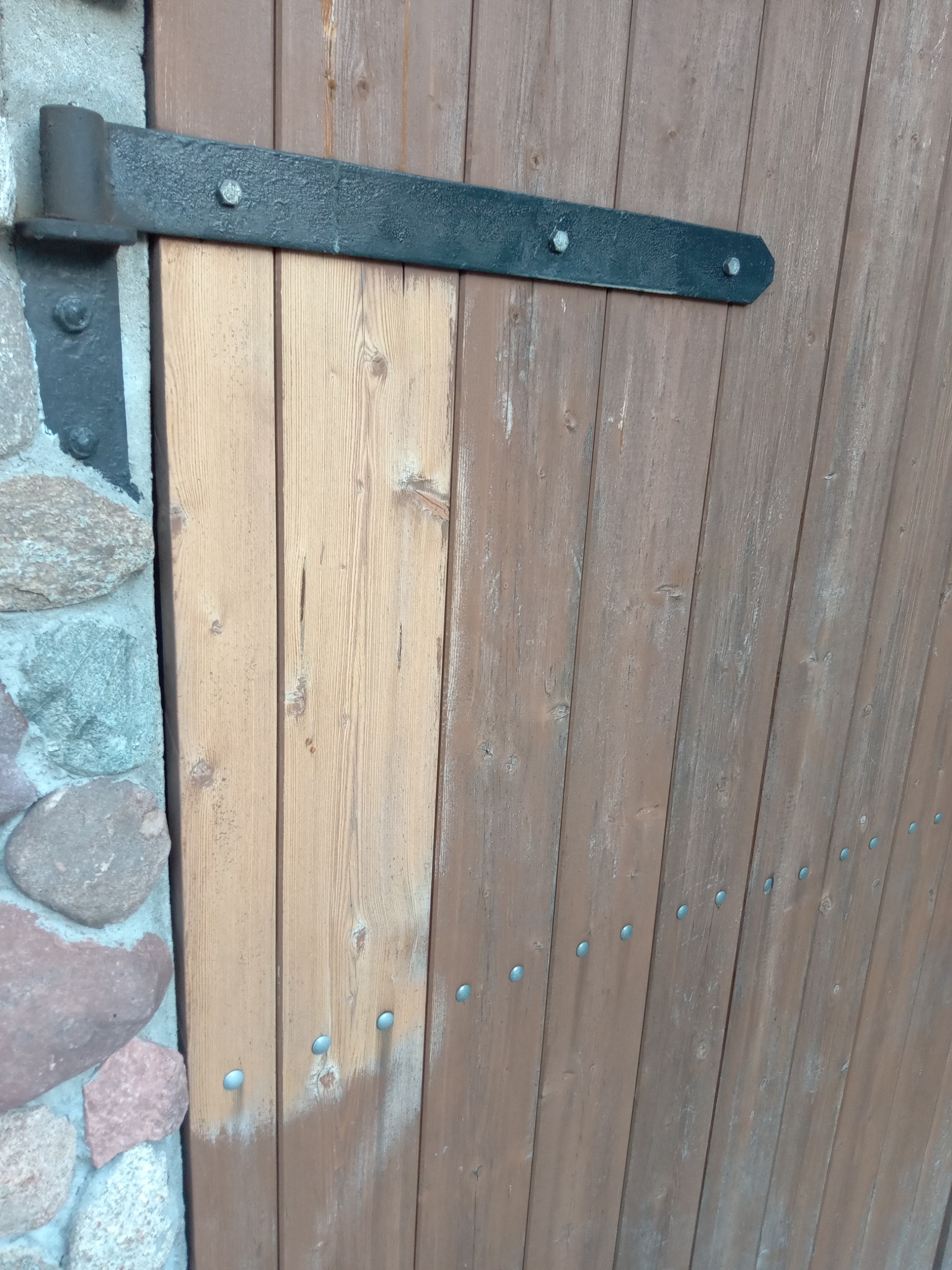 Fragment drewnianych drzwi z metalowymi okuciami i kamiennym murem po lewej stronie, widoczne ślady po malowaniu.