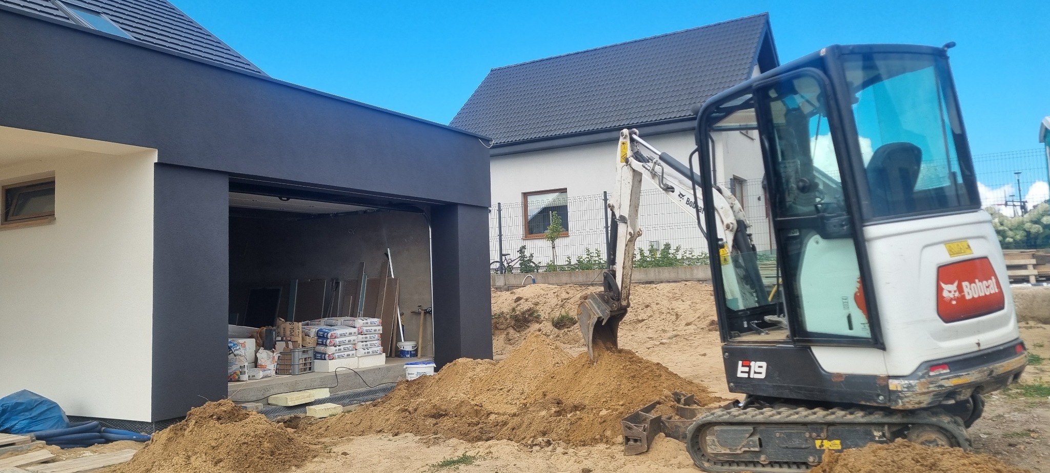 Mini koparka Bobcat E19 na placu budowy, z hałdą piasku przed budynkiem z czarno-białą elewacją. W tle dom jednorodzinny.