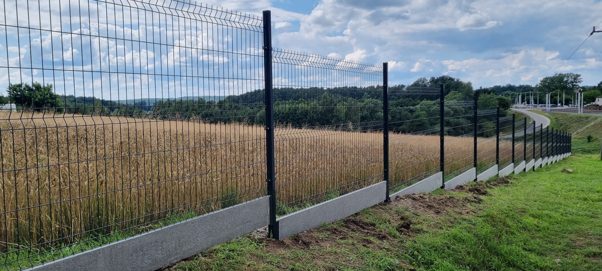 Nowoczesne ogrodzenie panelowe z podmurówką betonową, widok na pole zboża i błękitne niebo z chmurami. Perspektywa z boku, podkreślająca linię ogrodzenia.